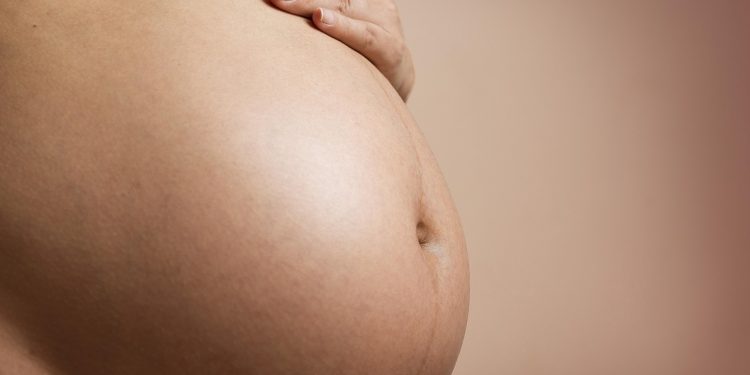 Kobieta w ciąży trzymająca się za brzuch dłonią, widoczny duży brzuch ciążowy.